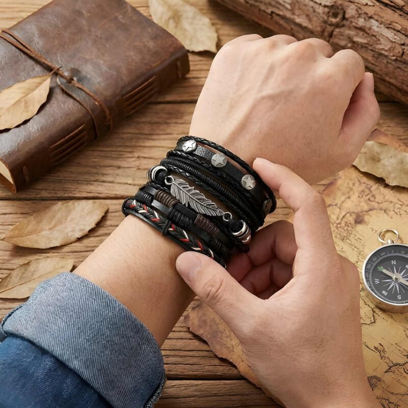 Man's wrist wearing a stacked black leather adventure bracelet set, styled on a wooden table with an antique compass, vintage leather journal, and dried autumn leaves.